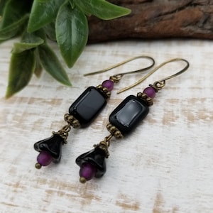 May include: A pair of drop earrings featuring a rectangular black bead, a small purple bead, and a black flower-shaped bead suspended from a bronze-toned metal hook. The components are connected by small bronze-toned metal spacers and wire links.