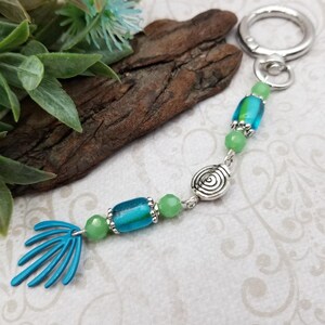 May include: A keychain featuring a silver-toned metal ring, a circular silver charm with a spiral design, two cylindrical blue and green glass beads, four small round light green beads, and a blue fan-shaped metal charm at the end.