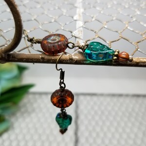 May include: Close-up of a handmade earring featuring a dark orange, round bead with a floral design, and a teal, heart-shaped bead. The earring is suspended from a wire hook and connected with small metal links.