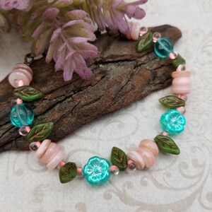 May include: A beaded bracelet featuring pink, green, and turquoise beads. The bracelet includes flower-shaped beads, leaf-shaped beads, and pink cylindrical beads. The bracelet is displayed on a piece of wood.