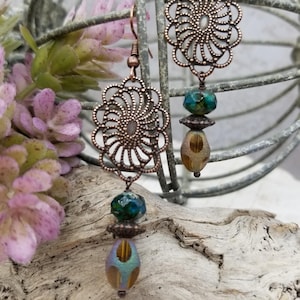 May include: Copper-colored filigree earrings with a floral design. Each earring features a dangling arrangement of teal, brown, and iridescent beads. The earrings are displayed against a backdrop of driftwood and faux flowers.