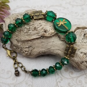 May include: A green beaded bracelet with a dragonfly charm. The bracelet features faceted green beads, antique brass-colored metal accents, and a round charm with a gold dragonfly design. The bracelet is approximately 7 inches long.