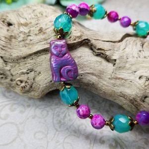 May include: A beaded bracelet featuring a purple cat charm. The bracelet has turquoise and magenta beads with gold-colored accents. The cat charm is carved and has a gradient of purple and blue hues. The bracelet is displayed on a piece of driftwood.