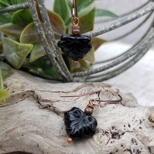 May include: A pair of dangle earrings featuring black carved leaf-shaped beads suspended from copper-toned metal hooks. Each earring includes a small copper-colored bead accent above the leaf, creating a cohesive design with a metallic finish.