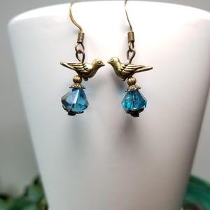 May include: A pair of earrings featuring small, bronze-colored bird charms perched above faceted, teardrop-shaped blue crystals. The earrings hang from bronze-colored ear wires, creating a delicate and whimsical design. The earrings are displayed against a white background.
