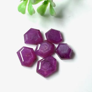 Puede incluir: Seis gemas hexagonales facetadas de color magenta intenso. Las gemas están dispuestas sobre una superficie blanca, con una ramita de flores artificiales verdes y blancas en el fondo. Las gemas tienen una superficie pulida y reflectante.