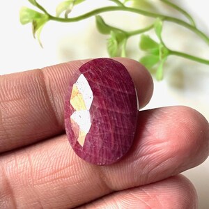 Op de afbeelding: Een gefacetteerde, ovaalvormige robijn edelsteen. De robijn heeft een dieprode kleur met een gepolijst oppervlak. De edelsteen wordt in een hand gehouden, met een takje groene bladeren op de achtergrond.