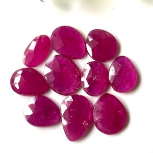 May include: A collection of faceted, oval-shaped, and teardrop-shaped ruby gemstones. The gemstones are a deep, vibrant pinkish-red color and are arranged on a white surface. The gems have a polished, reflective surface.