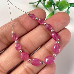 Può includere: Un filo di pietre preziose ovali rosa infilate su un filo d'argento. Le pietre preziose sono traslucide e hanno un leggero luccichio.