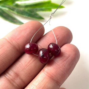 Puede incluir: Tres cuentas de piedras preciosas de color rojo oscuro, pulidas y ensartadas en un fino alambre de plata. Las cuentas son ovaladas y tienen un acabado brillante. Fondo blanco con follaje verde.