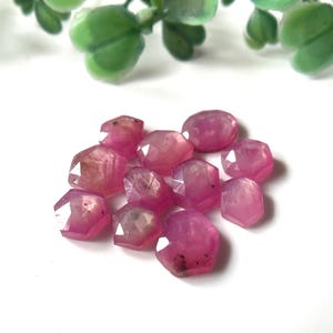 May include: A group of ten pink faceted gemstone beads. The beads are all different shapes and sizes, but they are all cut in a similar style. The beads are arranged in a loose pile on a white surface.