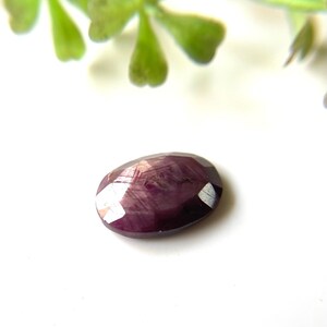 Op de afbeelding: Een gefacetteerde, ovaalvormige, dieprode edelsteen met witte reflecterende facetten. De edelsteen ligt tegen een witte achtergrond, met een vage groene plant in de linkerbovenhoek. De steen is waarschijnlijk een robijn.