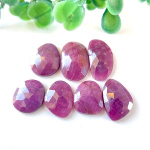 May include: Seven faceted pink ruby gemstones, each with a unique shape and cut, arranged in a row on a white background.