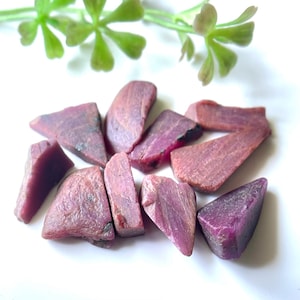 Può includere: Una collezione di dieci pietre grezze di rubino rosa. Le pietre grezze sono di varie forme e dimensioni, alcune sono triangolari e altre più arrotondate.