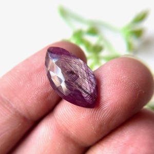 Op de afbeelding: Een gefacetteerde paarse edelsteen met een marquise-vorm. De edelsteen heeft een zilveren glans en wordt in een hand gehouden.