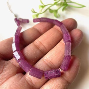 Może przedstawiać: Bransoletka z fasetowanych, prostokątnych, różowo-fioletowych kamieni szlachetnych. Kamienie szlachetne są nawleczone na bransoletkę. Bransoletka jest trzymana w dłoni, z białym tłem i zielonymi gałązkami w tle.