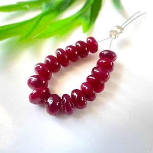 May include: A strand of polished, oval-shaped ruby beads. The beads are a deep, rich red color and are strung together with a silver-colored wire. The beads are approximately 0.25 inches in length. The background is white with green leaves.