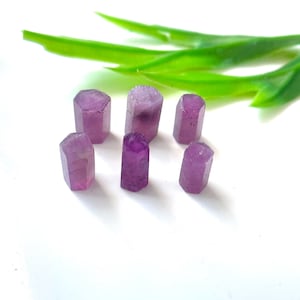 May include: Seven small, faceted, purple-pink gemstone crystals are arranged on a white surface. The crystals are in various sizes and shapes, with a hexagonal or rectangular form. Green, leaf-like accents are in the background.