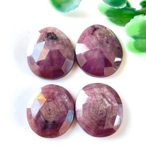 Peut inclure: Quatre cabochons de pierres précieuses facettées roses. Les pierres précieuses sont rondes et ont une forme légèrement irrégulière. Elles ont une couleur rose avec une légère teinte violette. Les pierres précieuses sont posées sur un fond blanc.