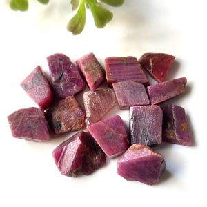 May include: A collection of rough, pink ruby gemstones. The stones are various sizes and shapes, with some having a more rectangular shape and others being more irregular.