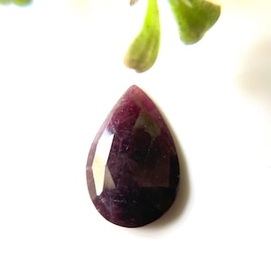 Puede incluir: Una piedra preciosa de color púrpura oscuro en forma de pera con un corte facetado. La piedra preciosa está sobre un fondo blanco.