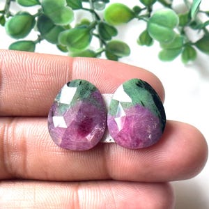 May include: Two faceted, teardrop-shaped gemstones. The gems feature a mix of pink, green, and clear facets. The gemstones are held between fingers, with a blurred green plant in the background.