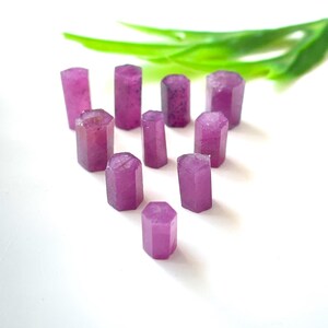 May include: A collection of raw, hexagonal-shaped ruby crystals in varying sizes. The crystals are a deep pinkish-red color, with a slightly translucent appearance. Green leaves are in the background, set against a white surface.