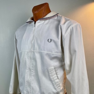 Może przedstawiać: Biała kurtka zapinana na zamek błyskawiczny z białym, żebrowanym kołnierzem i mankietami. Kurtka ma niebiesko-czerwony pasek na kołnierzu i mankietach. Kurtka ma małe haftowane logo na lewej piersi.