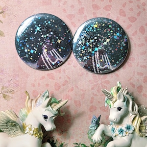 May include: Two circular pin-back buttons featuring a dark, starry night sky design with holographic star accents. Below the buttons are two white unicorn figurines with iridescent manes, gold horns, and floral neck garlands on a pink background.