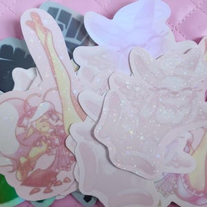 May include: A collection of pastel-colored stickers with glitter accents. The stickers feature illustrations of a character in a dress and hat, and other abstract shapes. The stickers are arranged on a pink surface.