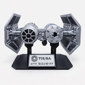Könnte beinhalten: Silberfarbenes TIE/SA-Modellraumschiff auf einem schwarzen Ständer. Das Modell hat zwei kugelförmige Cockpits, die durch eine zentrale Struktur verbunden sind, mit abgewinkelten Sonnenkollektoren, die sich von jeder Seite erstrecken. Die Basis enthält eine Plakette mit Text und einem Symbol.