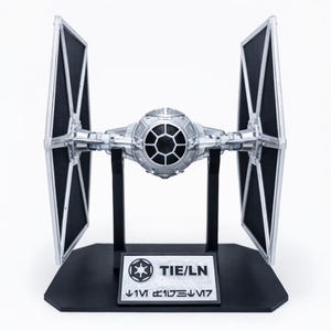 Könnte beinhalten: Silbernes TIE/LN Star Wars Modell mit schwarzen Solarpanelen, auf schwarzem Sockel. Der Sockel trägt die Aufschrift "TIE/LN" und Symbole. Detaillierte Nachbildung des ikonischen Raumschiffs, ein Sammlerstück.