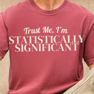 Può includere: Una t-shirt a maniche corte con girocollo in un colore rosso mattone tenue. La parte anteriore del capo presenta il testo "Trust Me, I'm STATISTICALLY SIGNIFICANT" stampato in un grande carattere serif color crema sulla zona del petto.