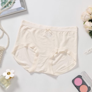 May include: A pair of solid cream-colored seamless underwear featuring a high-waisted design with a small, delicate bow centered at the waistband. The fabric appears smooth and lightweight, providing full coverage with clean, finished edges.