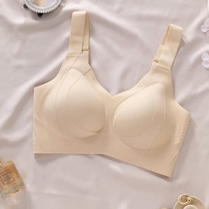 May include: A beige wireless bra featuring wide shoulder straps with adjustable sliders, molded cups with a seamless construction, and a deep V-neckline. The garment is crafted from a smooth, solid-colored fabric with subtle contouring lines.