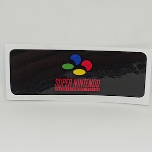 May include: A rectangular sticker featuring the Super Nintendo Entertainment System logo. The logo includes a black background with the words "SUPER NINTENDO ENTERTAINMENT SYSTEM" in red. The logo also has four colored shapes: blue, green, yellow, and red.