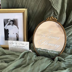 Puede incluir: Una exhibici&oacute;n conmemorativa con una foto en blanco y negro enmarcada de un hombre con uniforme naval, un espejo con marco dorado con las palabras "In Loving Memory" y una tarjeta con el nombre. Los art&iacute;culos est&aacute;n dispuestos sobre una tela verde salvia drapeada.