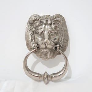 Puede incluir: Un aldaba de puerta con cabeza de león plateada con un asa de anillo. La cara del león está detallada con una melena, ojos, nariz y boca. El asa de anillo está unida debajo de la cara del león.