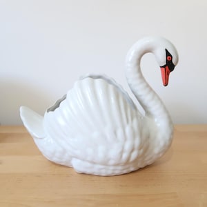 May include: A white ceramic swan-shaped planter with a curved neck and a red and black beak. The swan has detailed feather textures and a hollowed-out back for planting flowers or displaying items. The planter sits on a wooden surface.