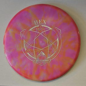 May include: Axiom Discs Fission Hex midrange driver disc. The disc features a pink and orange tie-dye pattern. The disc has the word "HEX" and "MIDRANGE DRIVER" printed on it, along with a geometric design and the word "FISSION".