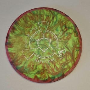 May include: A green, red, and yellow disc golf disc with a white design. The disc has the words "Rhythm" and "Innova Discs" printed on it.