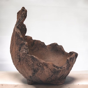 May include: A brown, hand-formed ceramic bowl with a rough, textured exterior and an uneven, jagged rim. The vessel features a deep, curved basin and sits on small, integrated feet, showcasing a dark, earthy finish with mottled surface details.