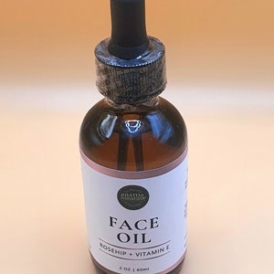 May include: A glass bottle of face oil with a dropper top. The bottle is labelled "Rayo Naturale Face Oil Rosehip + Vitamin E 2 oz | 60 ml".