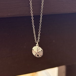 May include: A silver necklace with a small sand dollar pendant. The necklace is hanging on a wooden railing.