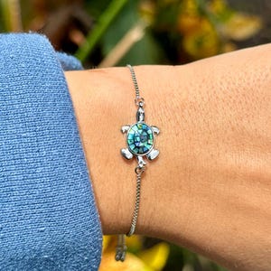 May include: A silver bracelet featuring a turtle charm with a blue and green iridescent shell. The bracelet is worn on a wrist, with a blue long-sleeved shirt in the background. The charm is approximately 0.75 inches in size.