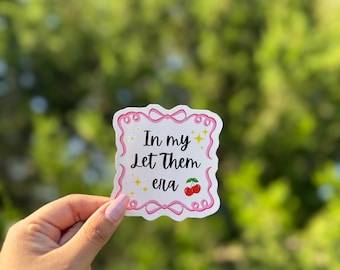 May include: A white sticker with pink trim and the words "In my Let Them era" in black script. The sticker features yellow stars and a pair of red cherries. The sticker is held in front of a blurred green background.