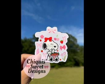 May include: A white sticker featuring Snoopy holding strawberries, adorned with red bows, flowers, and cherries. The sticker has the text "Chiquis Sweet Designs" in pink. The background is a blue sky and green grass.