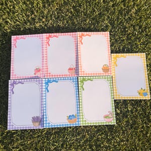 May include: Seven rectangular memo pads featuring a white center with a scalloped border, surrounded by a colored gingham frame. Each pad displays a small illustration of a basket containing items like fruit, flowers, or herbs, and a matching bow.