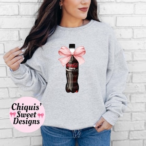 May include: A gray sweatshirt with a pink bow on a Coca-Cola Zero bottle. The text "Chiquis' Sweet Designs" is printed on the bottom left corner of the sweatshirt.