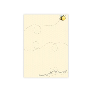 Puede incluir: Papel de carta amarillo claro con un patrón de panal y una abeja de dibujos animados. La abeja es amarilla y negra, con una cara sonriente. Una línea de puntos muestra la trayectoria de vuelo de la abeja. El texto "Buzzin' By With a Note From Maya" está en la parte inferior.